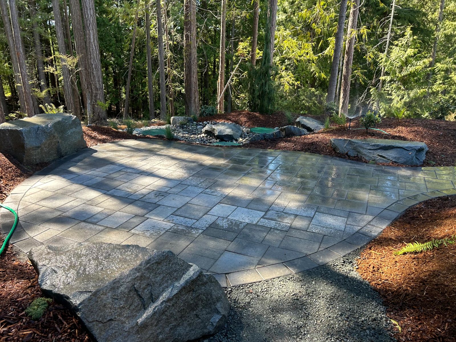 Wet paver patio framed by boulders and evergreens